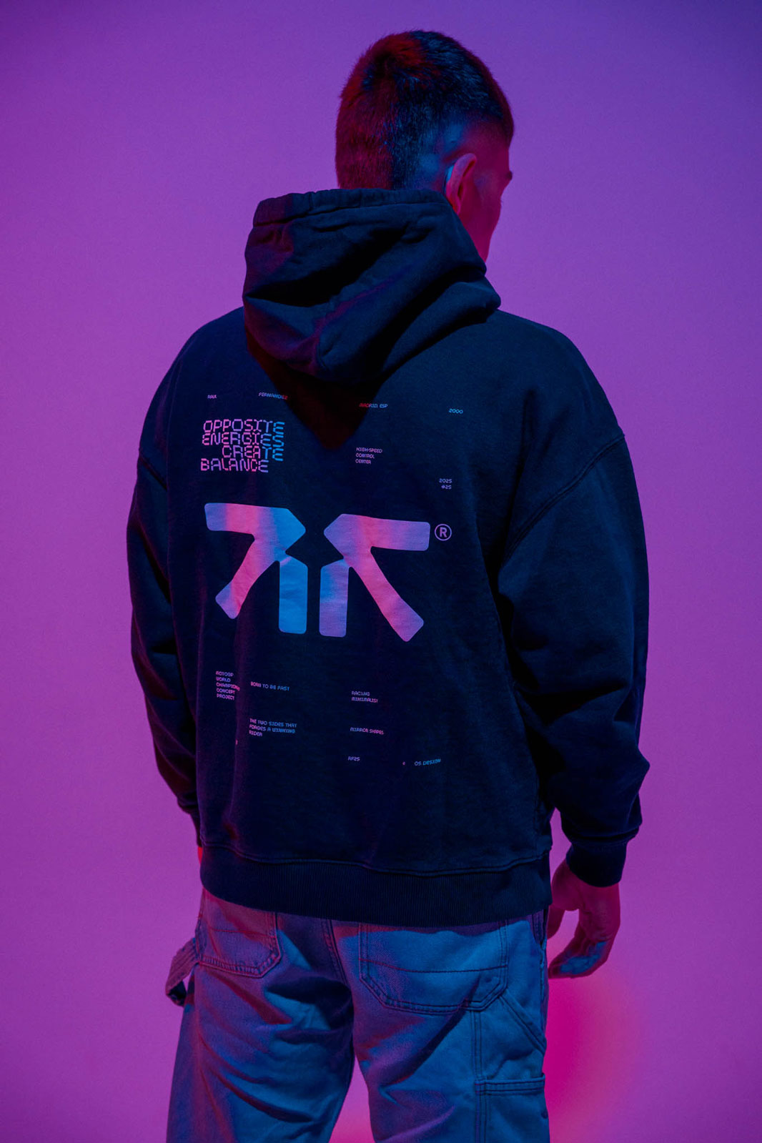 Person seen from behind wearing a black hoodie with colorful graphic text and symbols on the back, standing against a purple-lit background.