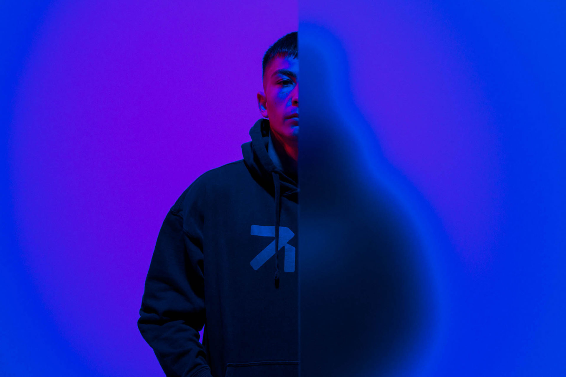 Young man wearing a black hoodie with an arrow symbol, half of his face obscured by a blue translucent panel against a purple and blue background.