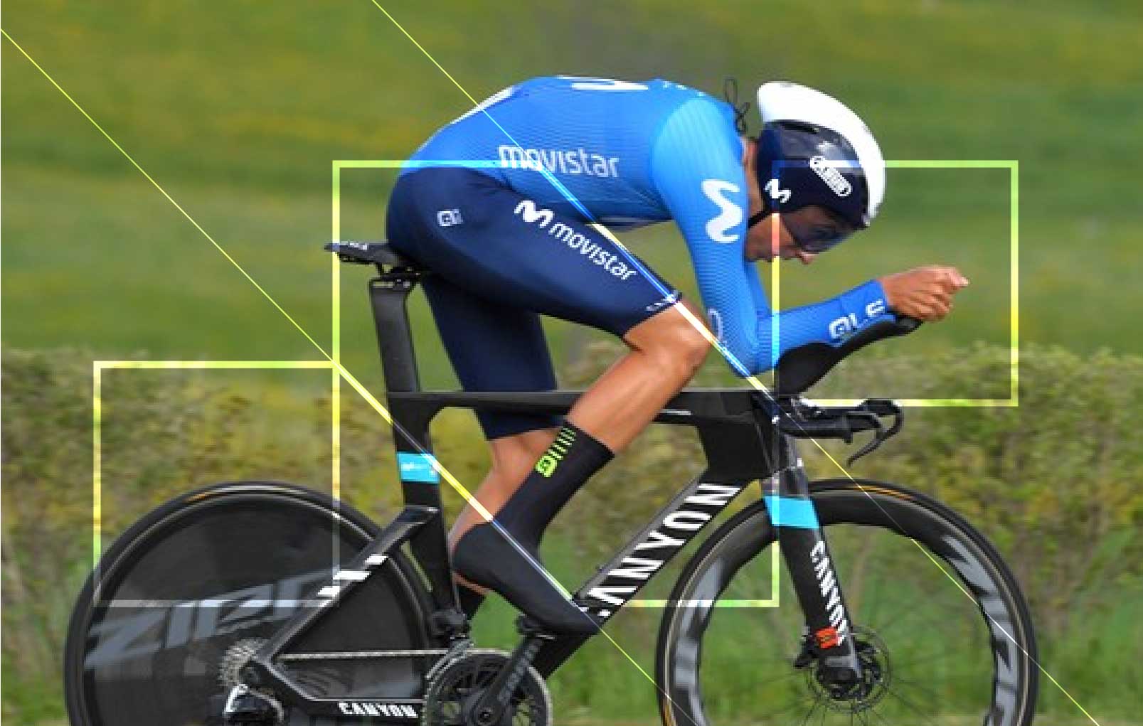 Cyclist in blue Movistar kit riding a black Canyon time trial bike with aerodynamic helmet on a green background.