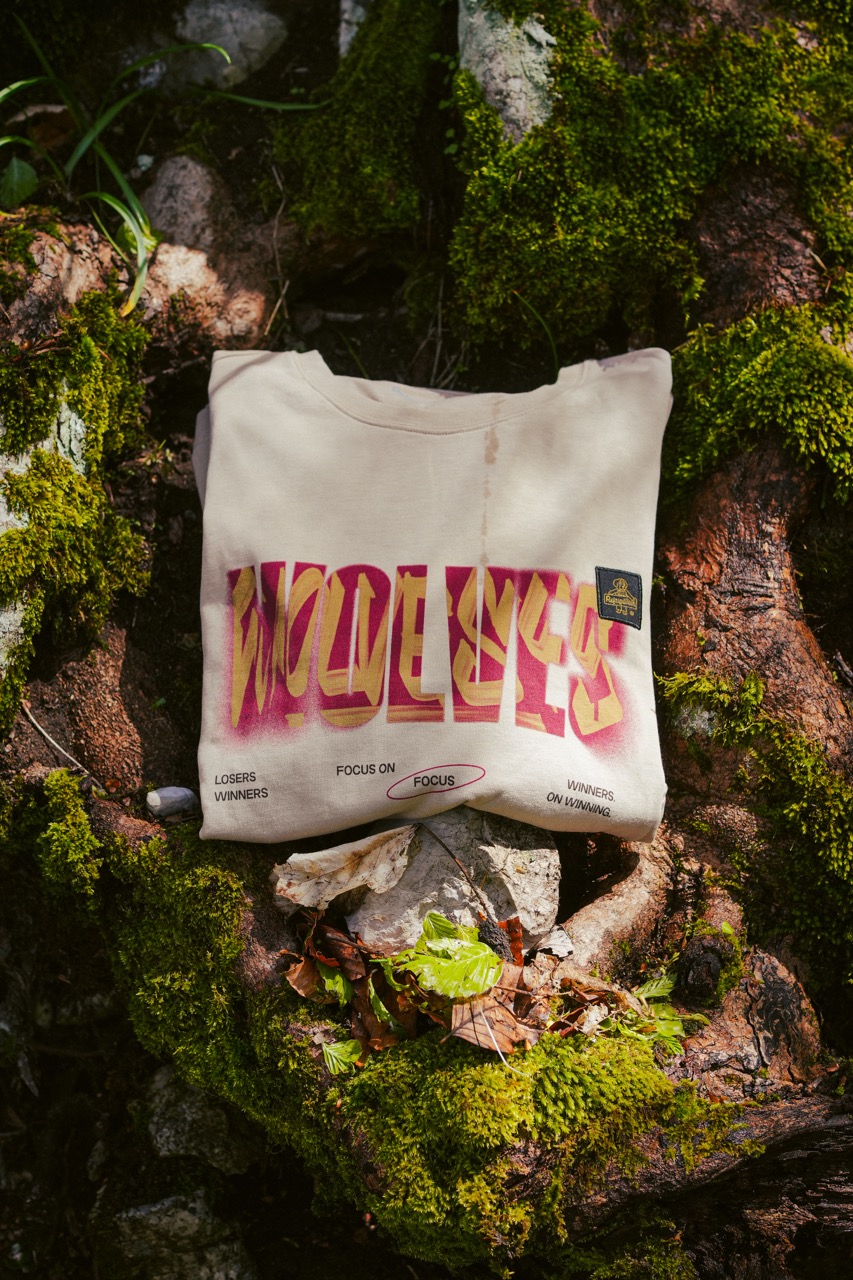 Folded cream-colored sweatshirt with bold multicolor text reading 'WOLVES' and smaller printed words 'LOSERS WINNERS', 'FOCUS ON FOCUS', and 'WINNERS ON WINNING' placed on moss-covered tree roots outdoors.