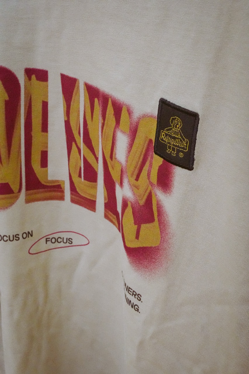 Close-up of cream-colored fabric with yellow and red graffiti-style text and a small brown patch featuring a yellow outlined dog and the word 'RefrigiWear'.