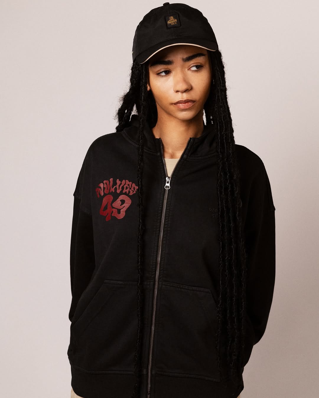 Person with long braided hair wearing a black zip-up hoodie with red 'Wolves 49' text and a black cap.