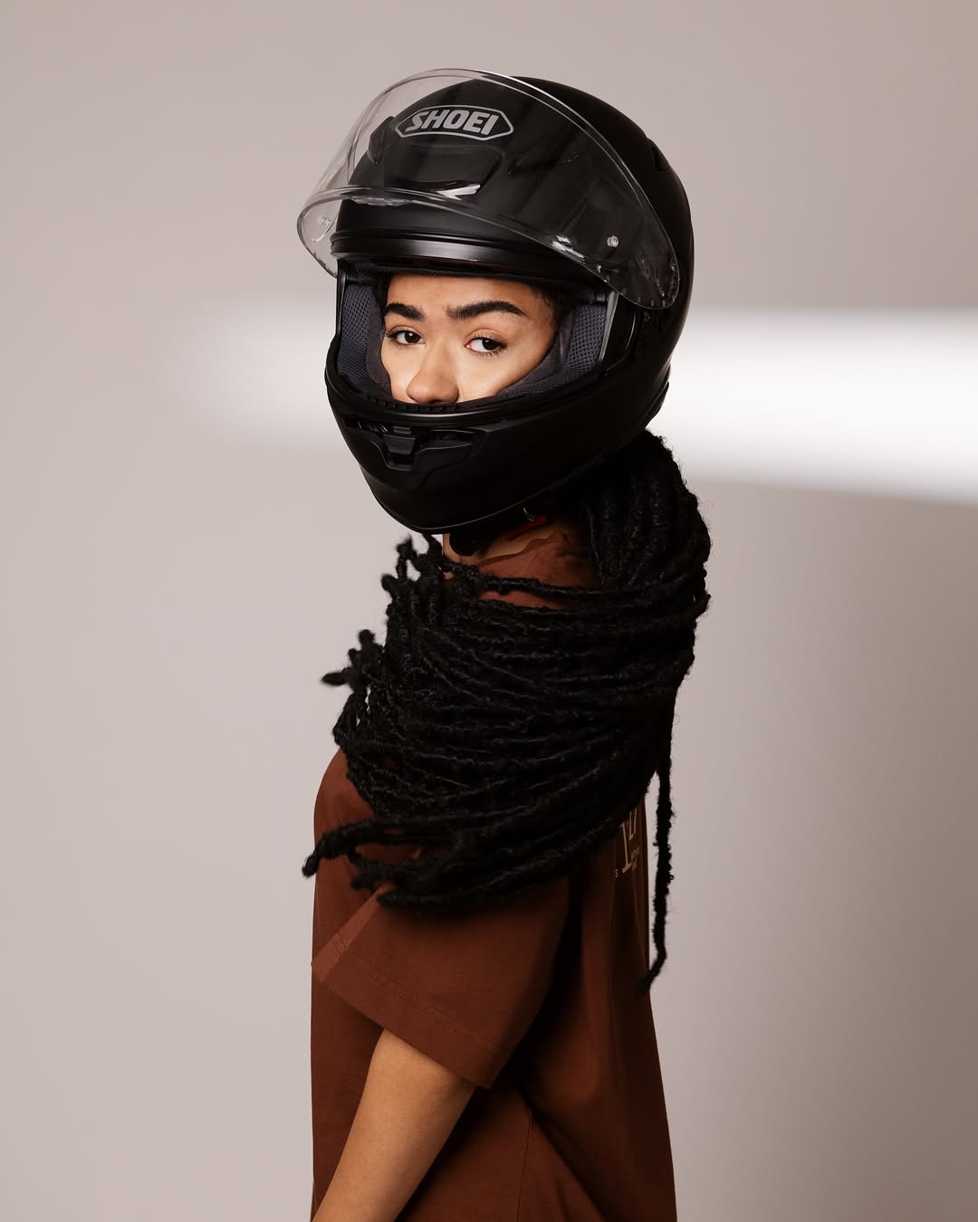 Person with long black dreadlocks wearing a black Shoei motorcycle helmet and a brown shirt against a neutral background.