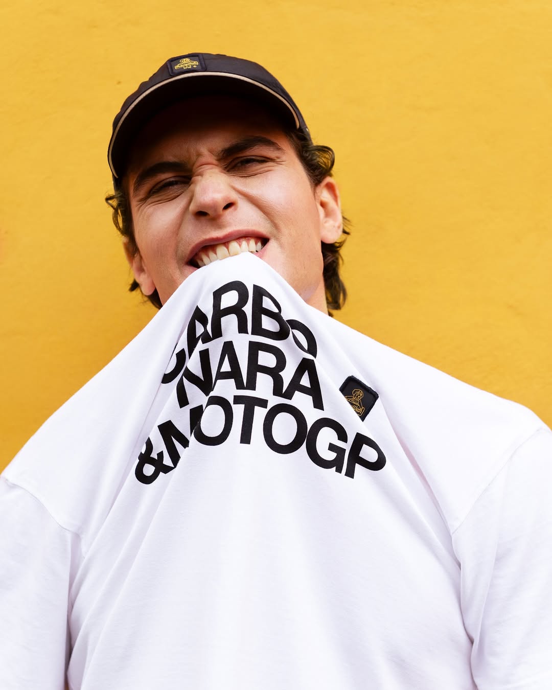 Young man wearing a black cap bites a white shirt with bold black text against a yellow background.