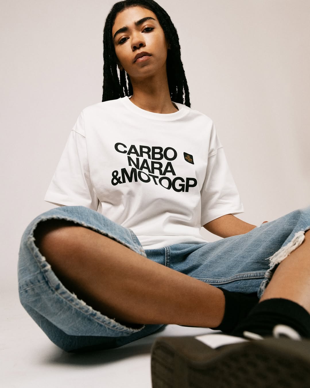 Person with long twisted hair wearing a white t-shirt with black text and ripped blue jeans, sitting cross-legged.