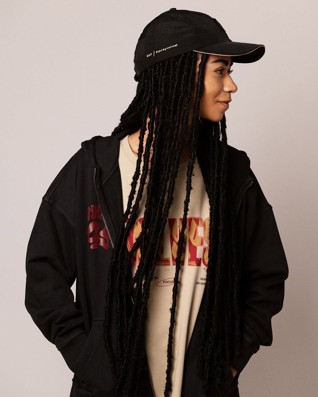 Person wearing a black cap, black zip-up hoodie, and beige graphic shirt with long black dreadlocks looking to the right.