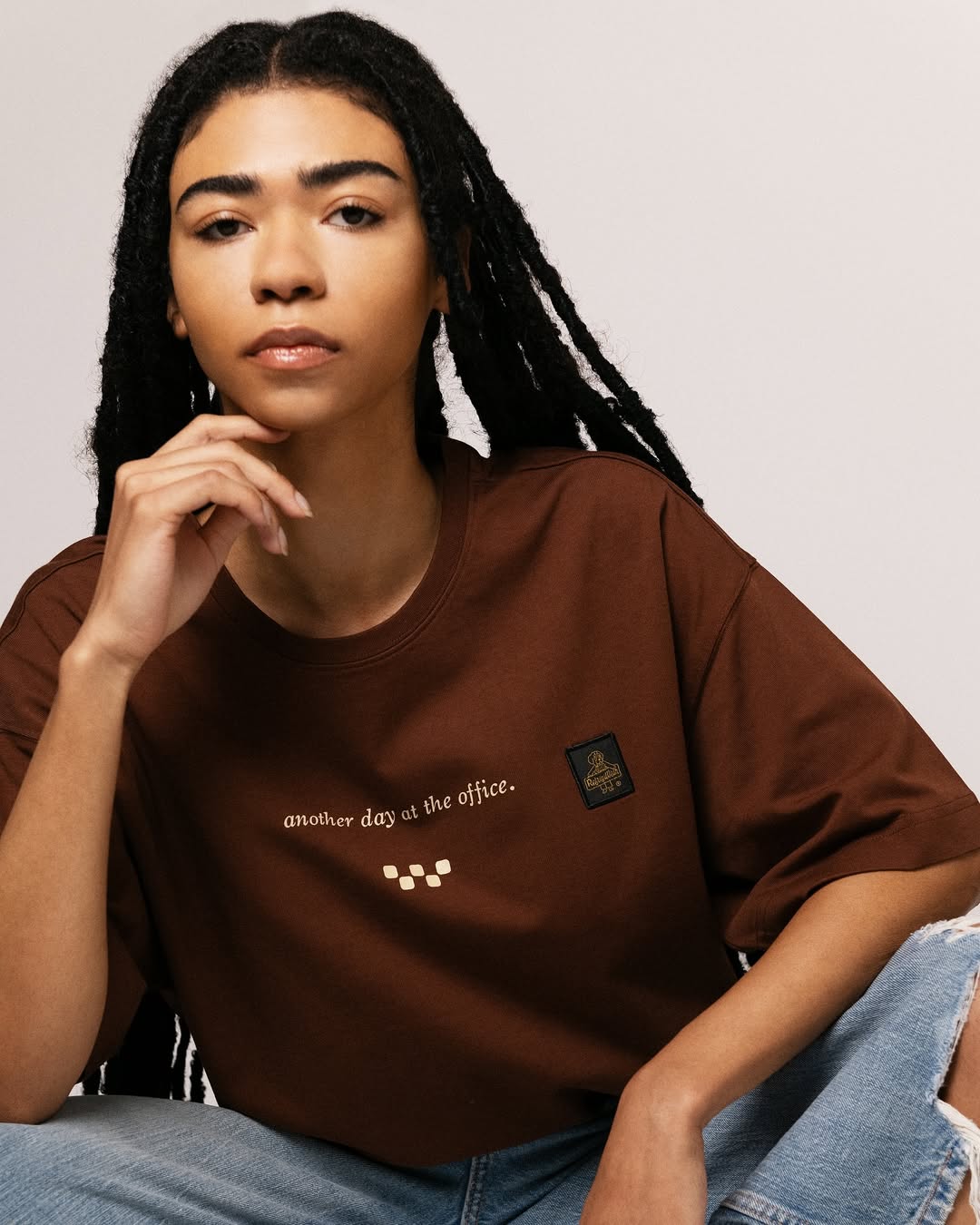 Person with long braided hair wearing a brown t-shirt reading 'another day at the office' and ripped light blue jeans.