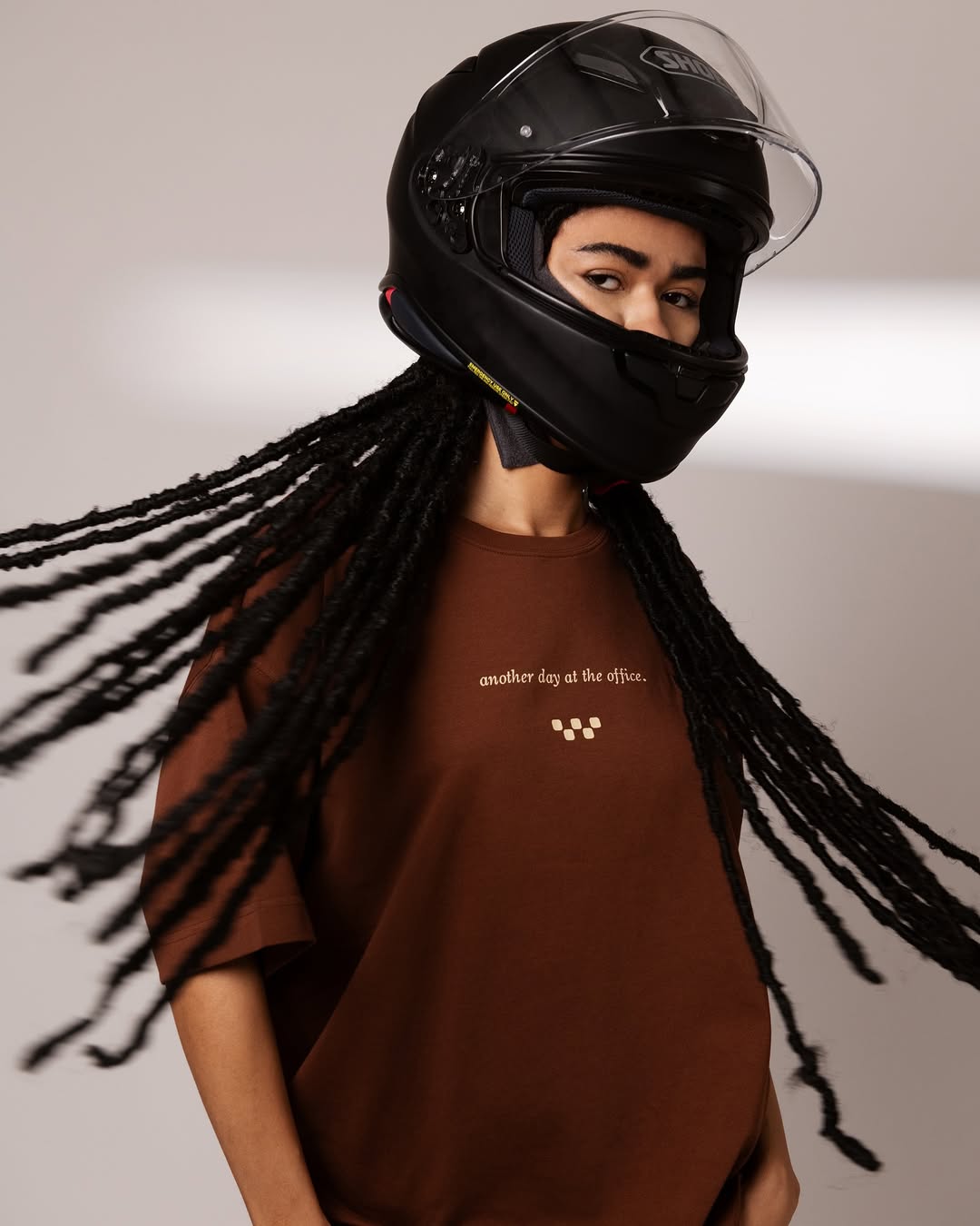 Person wearing a black motorcycle helmet and brown shirt with text 'another day at the office.' and long braided hair flowing.