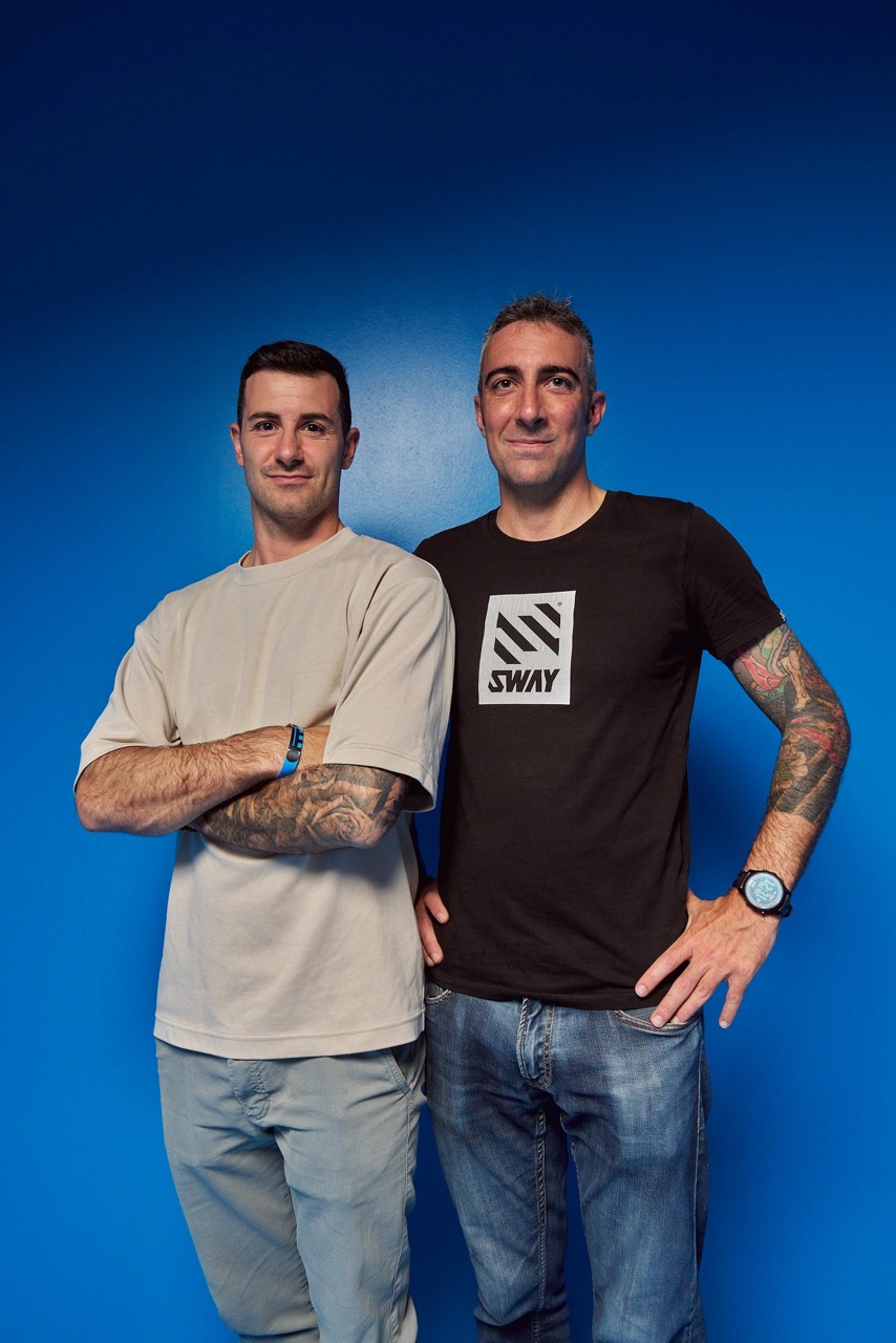 Two men with tattoos, one in a beige shirt and the other in a black shirt with a SWAY logo, standing against a blue wall.