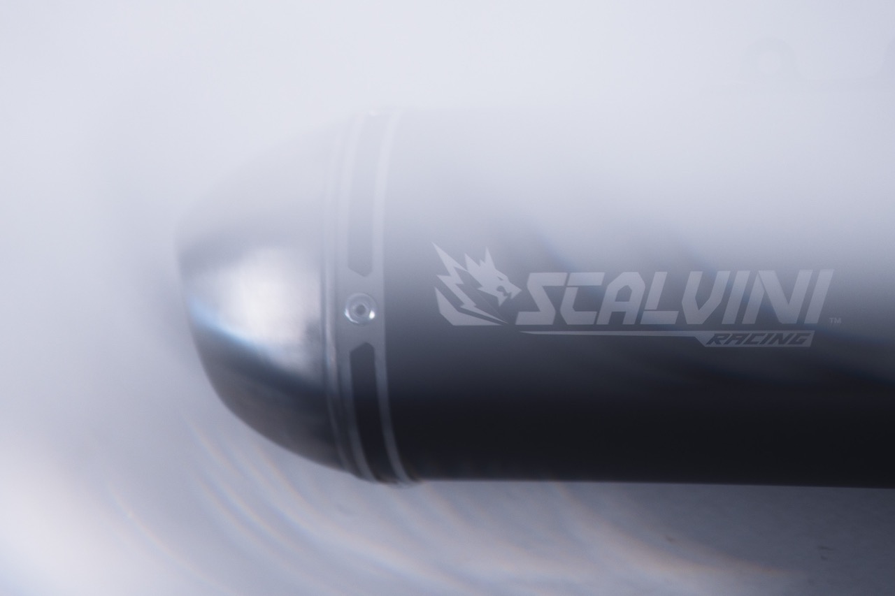 Close-up of a black cylindrical Scalvini Racing motorcycle exhaust pipe with a metallic tip, partially obscured by smoke or mist.