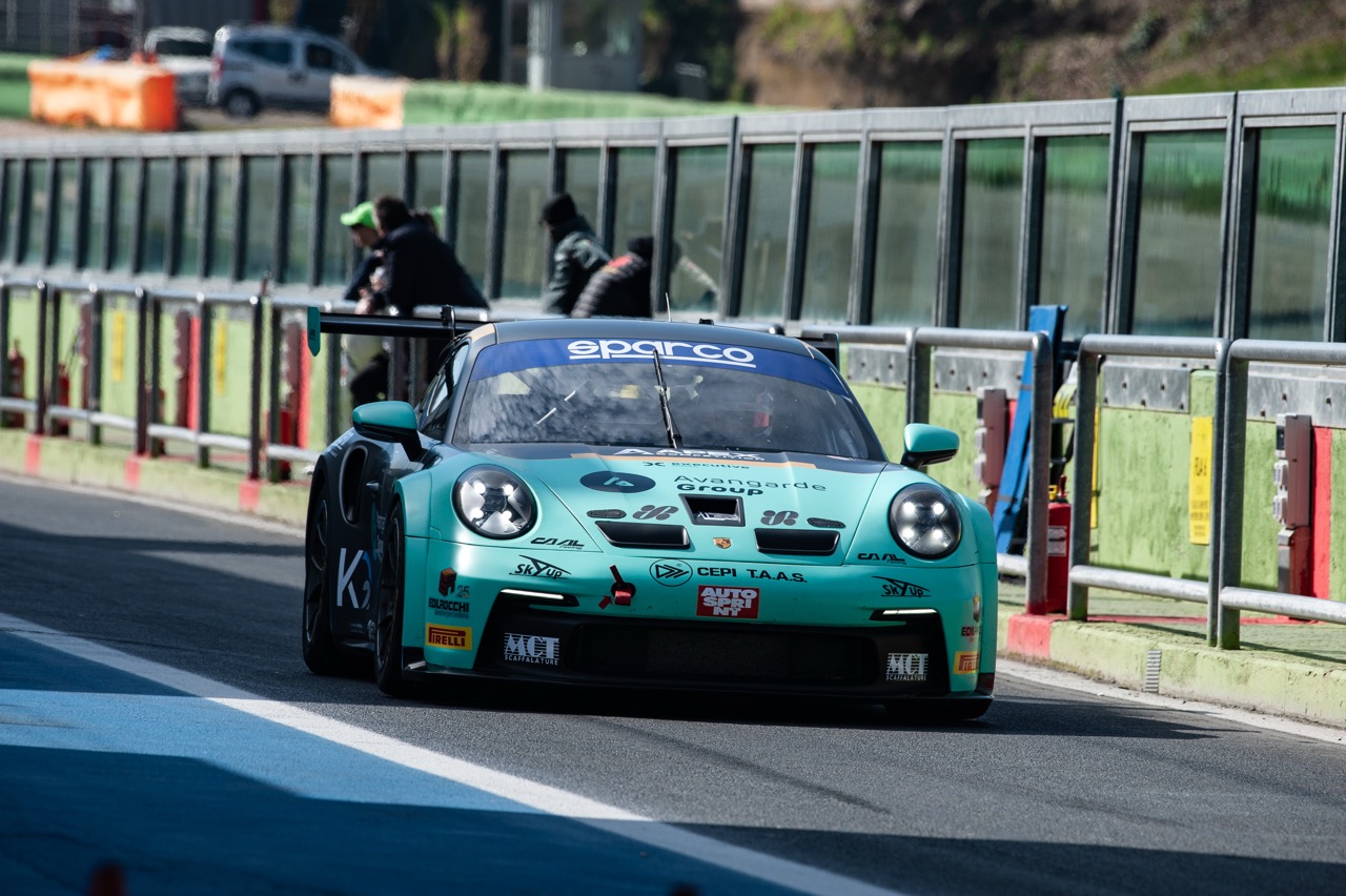 Front view of a teal Porsche race car with sponsor decals on a racetrack pit lane.