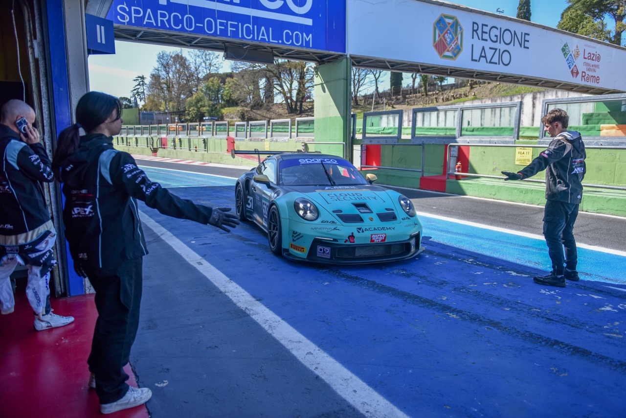 Teal and black race car entering pit lane with team members guiding on each side.