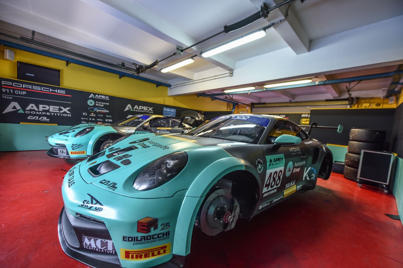 Two turquoise and black Porsche 911 Cup race cars in a garage, one without front wheels.