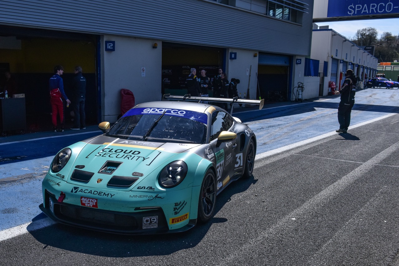 Light blue and black Porsche race car number 31 parked in a pit lane with mechanics and crew members nearby.
