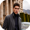 Young man wearing a dark coat and turtleneck standing outdoors near a white vehicle and classical building columns.
