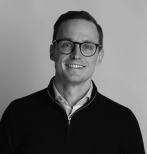 Smiling young man in a gray suit and white shirt standing outdoors in front of a house.