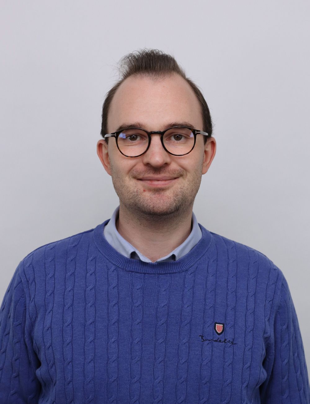 Smiling young man in a gray suit and white shirt standing outdoors in front of a house.
