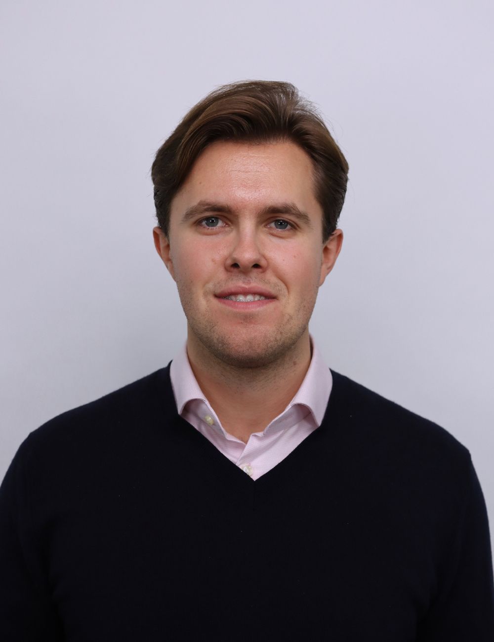 Smiling young man in a gray suit and white shirt standing outdoors in front of a house.