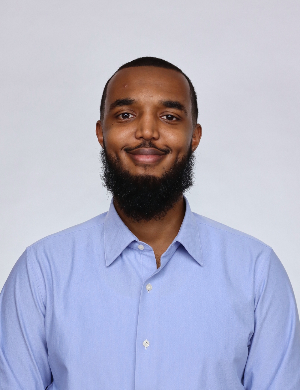 Smiling young man in a gray suit and white shirt standing outdoors in front of a house.