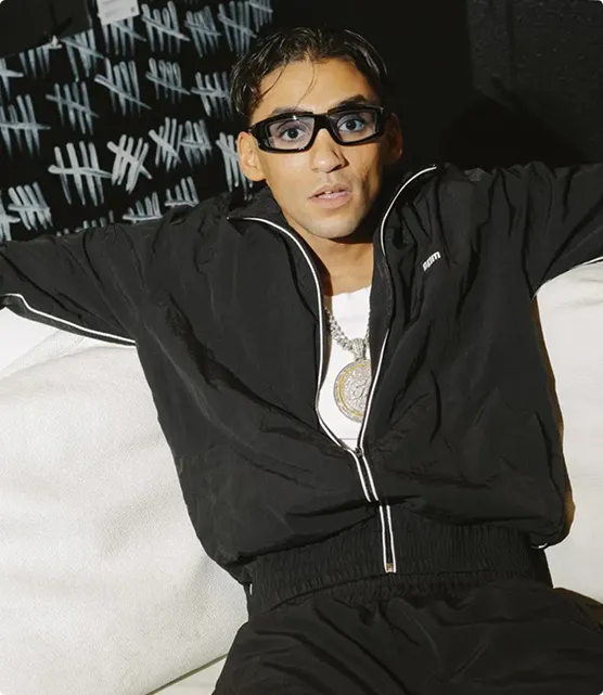 Mann in schwarzer Sportjacke und schwarzer Hose sitzt entspannt auf einem weißen Sofa vor einer schwarzen Wand mit weißen Strichmarkierungen.