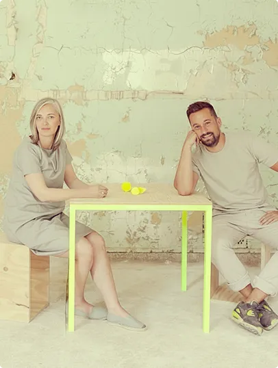 Frau und Mann in grauer Kleidung sitzen an einem hellgelb beleuchteten Tisch vor einer abblätternden Wand.