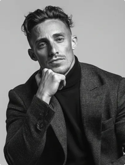 Schwarz-weiß-Porträt eines nachdenklichen Mannes mit gezwirbeltem Haar und leichtem Bart, der sein Kinn auf seine Faust stützt, bekleidet mit einem dunklen Rollkragenpullover und einem karierten Sakko.