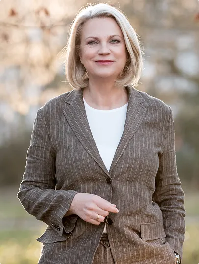 Lächelnde blonde Frau mittleren Alters in einem braunen gestreiften Anzug vor unscharfem Hintergrund im Freien.