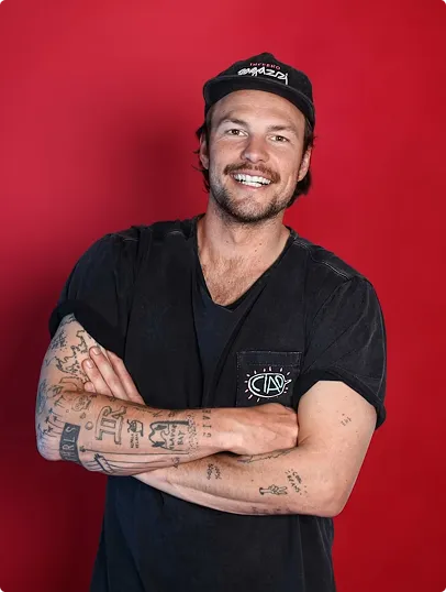 Lächelnder Mann mit schwarzem T-Shirt, Tattoos auf den Armen und schwarzer Kappe vor rotem Hintergrund.