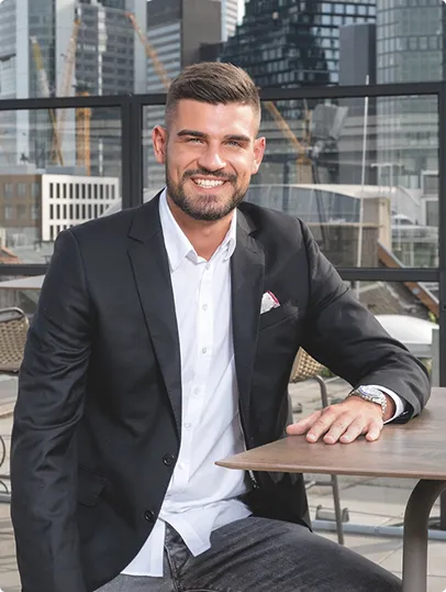 Junger Mann mit kurzem Haar und Bart in schwarzem Anzug und weißem Hemd sitzt draußen an einem Tisch mit Gebäuden im Hintergrund.