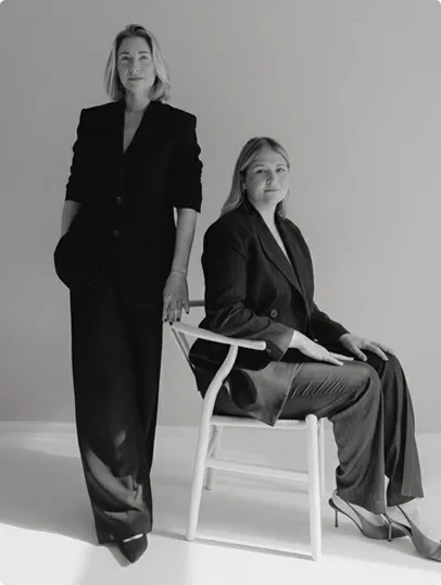 Zwei Frauen in schwarzen Anzügen posieren in einem minimalistischen Raum, eine steht und lehnt an einem Stuhl, die andere sitzt.