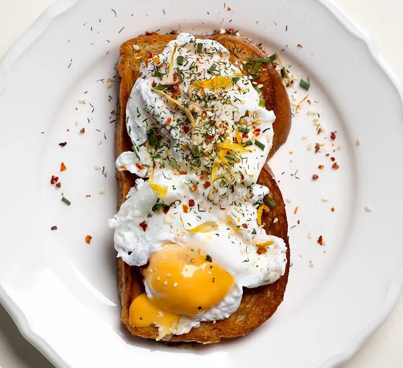 Toastbrot mit pochiertem Ei, garniert mit Kräutern, Sesam und Gewürzen auf einem weißen Teller.