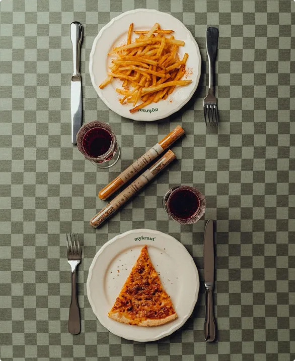 Zwei Teller auf einem karierten Tisch mit Pommes Frites auf dem oberen und einem Stück Pizza auf dem unteren Teller, jeweils mit Messer und Gabel, dazu zwei Gläser Rotwein und zwei Gewürzstreuer in der Mitte.
