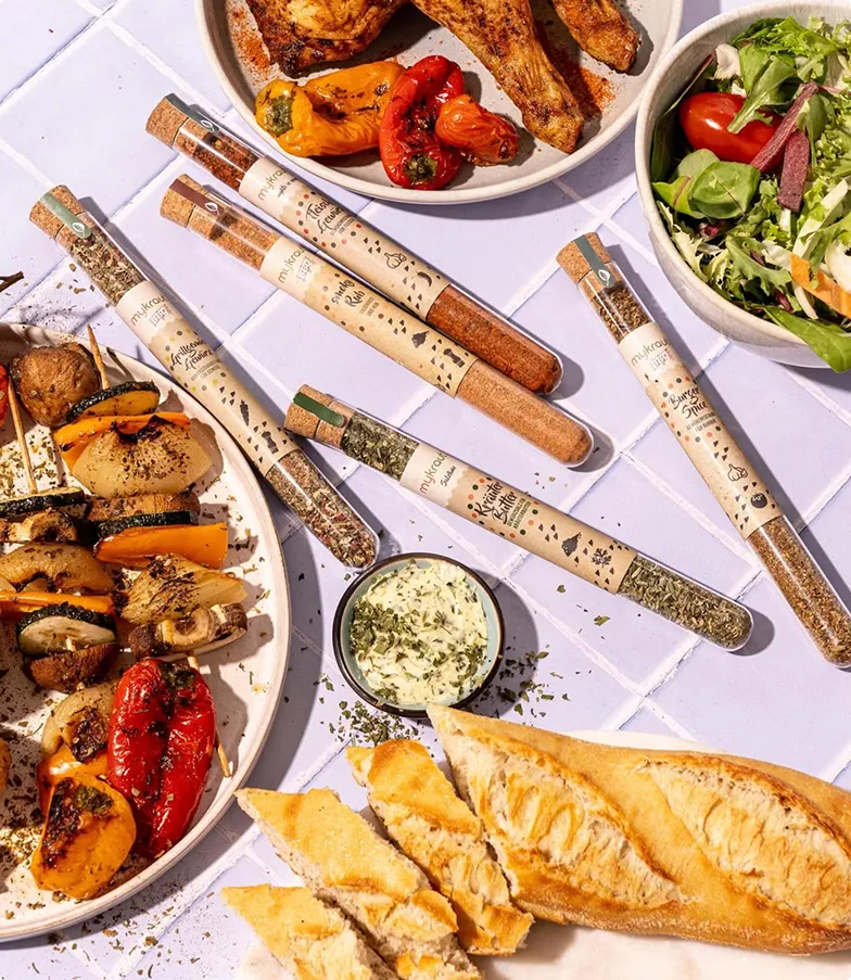 Grillteller mit gegrilltem Gemüse, Hühnchenschenkeln, Salat, Baguette und verschiedenen Gewürzröhrchen auf weißer Kacheloberfläche.