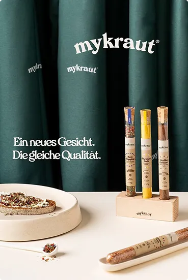 Drei Mykraut-Gewürzröhrchen in einem Holzhalter neben einem Brot mit Gewürzen auf einer weißen Fläche, vor grünem Vorhang mit Mykraut-Logo.