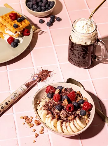 Schüssel mit Haferbrei, Bananenscheiben, Beeren und Granola neben Glas mit Schokolade und Sahne und Teller mit Waffeln und Beeren auf rosafarbenem Kachelboden.