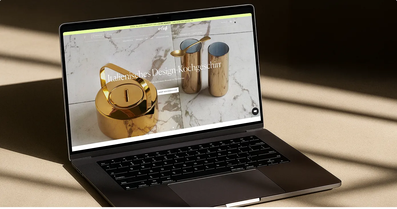 Laptop auf einem Tisch mit einer Website, die italienisches Design-Kochgeschirr zeigt, darunter ein goldener Teekessel und zwei goldene Becher mit einem Löffel.