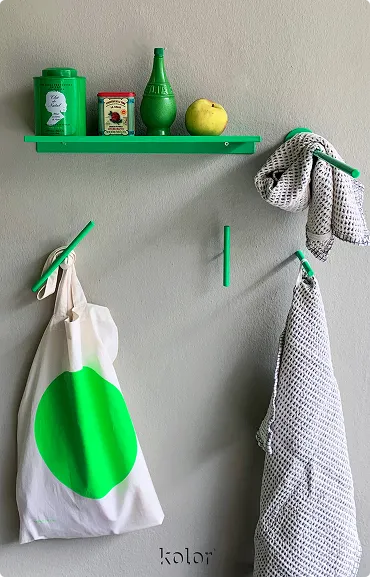 Grüne Wandablage mit drei Gegenständen und Apfel, darunter grüne Haken mit weißen Textilien und Tasche mit grünem Punkt.