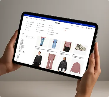 Zwei Hände halten ein Tablet, das eine Online-Shopping-Seite für Kleidung mit verschiedenen Artikeln wie Jeans, Pullover und Sneakern anzeigt.