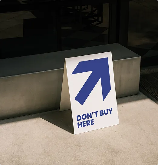 Ein Aufsteller mit einem großen blauen Pfeil nach oben rechts und dem Text 'DON'T BUY HERE' steht auf einem hellen Boden vor einer Betonbank.