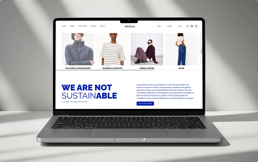 Laptop zeigt Website mit moderner Kleidungskollektion und großem Text 'WE ARE NOT SUSTAINABLE' auf weißem Hintergrund.