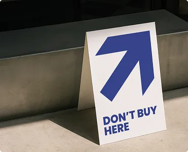 Schild mit einem blauen Pfeil nach rechts oben und dem Text 'DON'T BUY HERE' auf hellem Boden vor einer Treppenstufe.