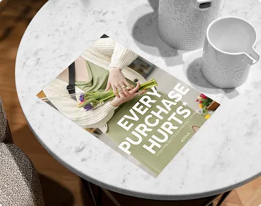 Flyer mit der Aufschrift 'Every Purchase Hurts' und einem Foto von Händen, die Blumen halten, auf einem runden weißen Marmor-Tisch neben zwei weißen Tassen.