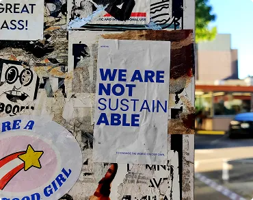 Plakat mit der Aufschrift 'WE ARE NOT SUSTAINABLE' an einer verzierten, wettergegerbten Wand im Freien.
