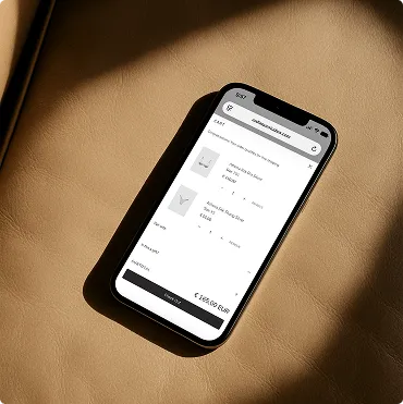 Smartphone auf beigem Lederuntergrund zeigt einen Online-Warenkorb mit zwei Schuhpaaren und Gesamtpreis in Euro.