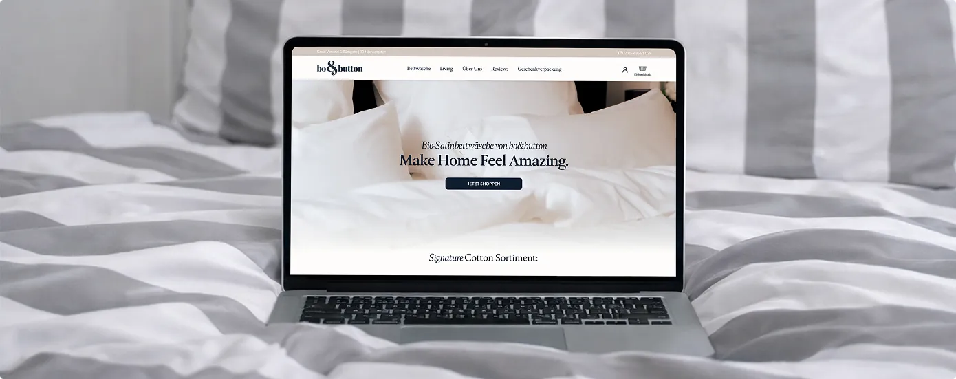 Laptop zeigt Webseite von bo&button für Bio-Satinbettwäsche, steht auf einem Bett mit grau-weiß gestreifter Bettwäsche.