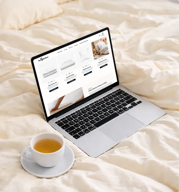 Laptop mit einem Online-Shop für Bettwaren auf einem cremefarbenen Bett, daneben eine Tasse Tee auf einem Unterteller.