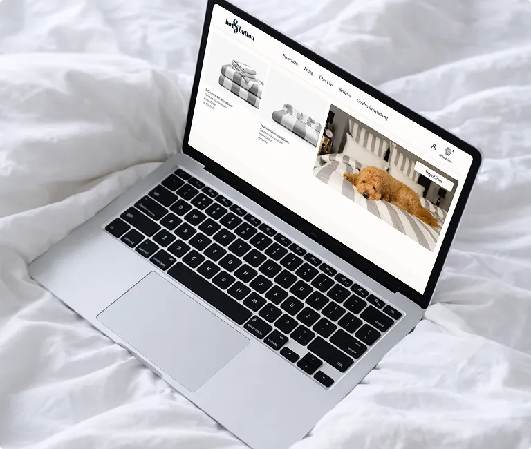 Laptop auf weißer Bettdecke zeigt Webseite mit Haustierbettprodukten und Bild eines Hundes auf gestreiftem Bett.