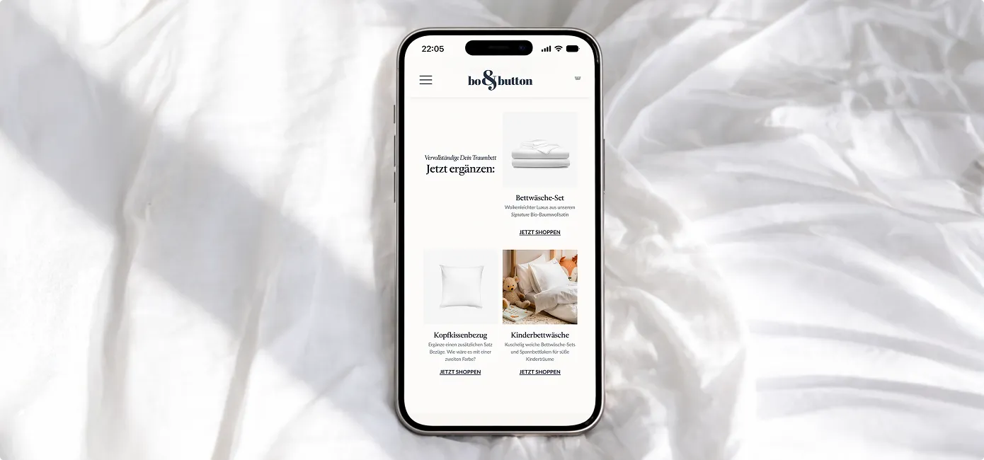 Smartphone auf weißem Bett mit geöffneter bo&button Webseite, die Bettwäsche, Kopfkissenbezug und Kinderbettwäsche zum Shoppen zeigt.