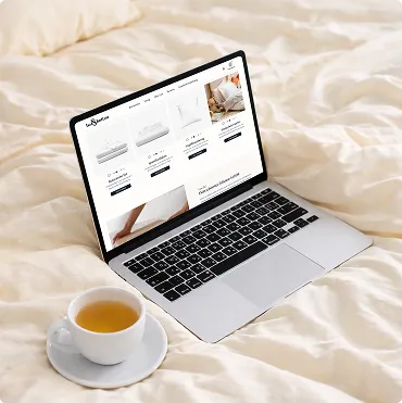 Laptop auf einem beigen Bett mit geöffnetem Online-Shop und einer Tasse Tee auf einer Untertasse daneben.