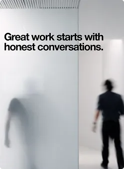 Text auf einer Wand: 'Great work starts with honest conversations.' Im Vordergrund sind zwei unscharfe Personen zu sehen.